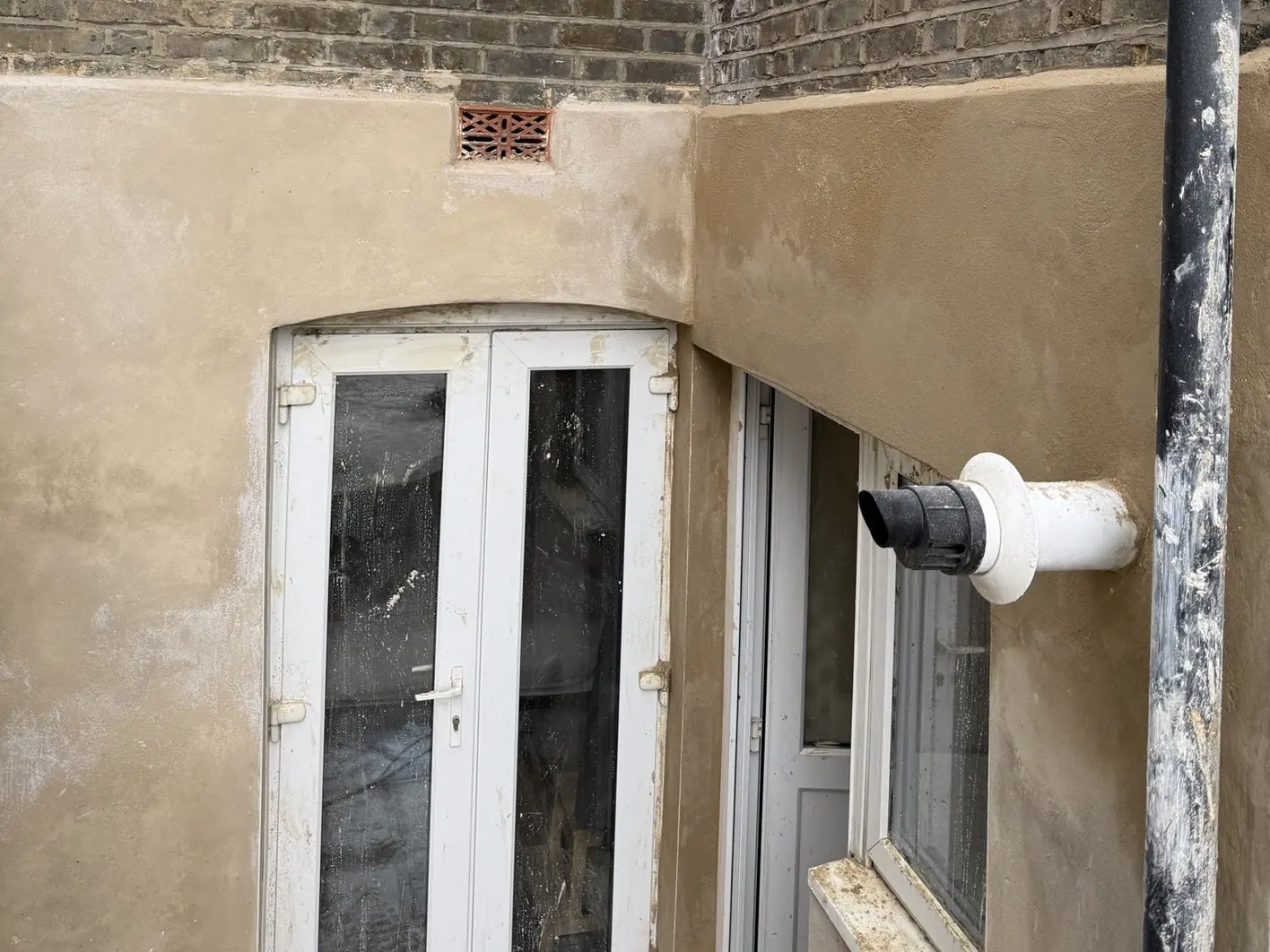 Rear elevation with fresh sand-coloured lime render across the lower walls, an arched door head, a terracotta airbrick set into the render and a flue terminal beside the downpipe