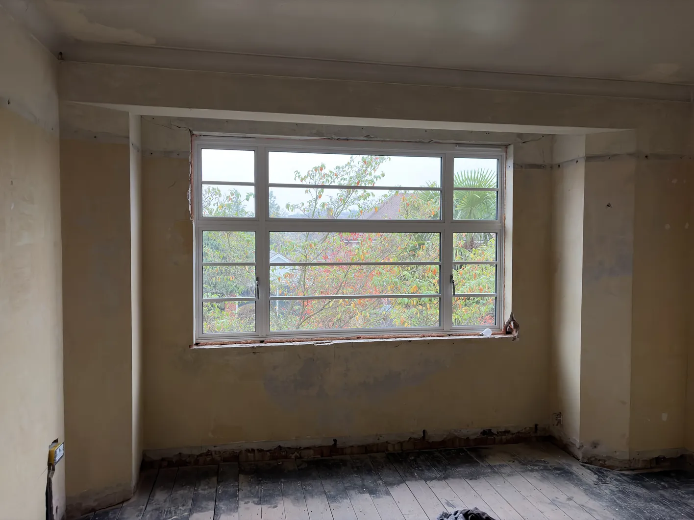 Interior of a room during renovation showing a large multi-pane window set into a deep reveal with damaged plasterwork and exposed timber floor
