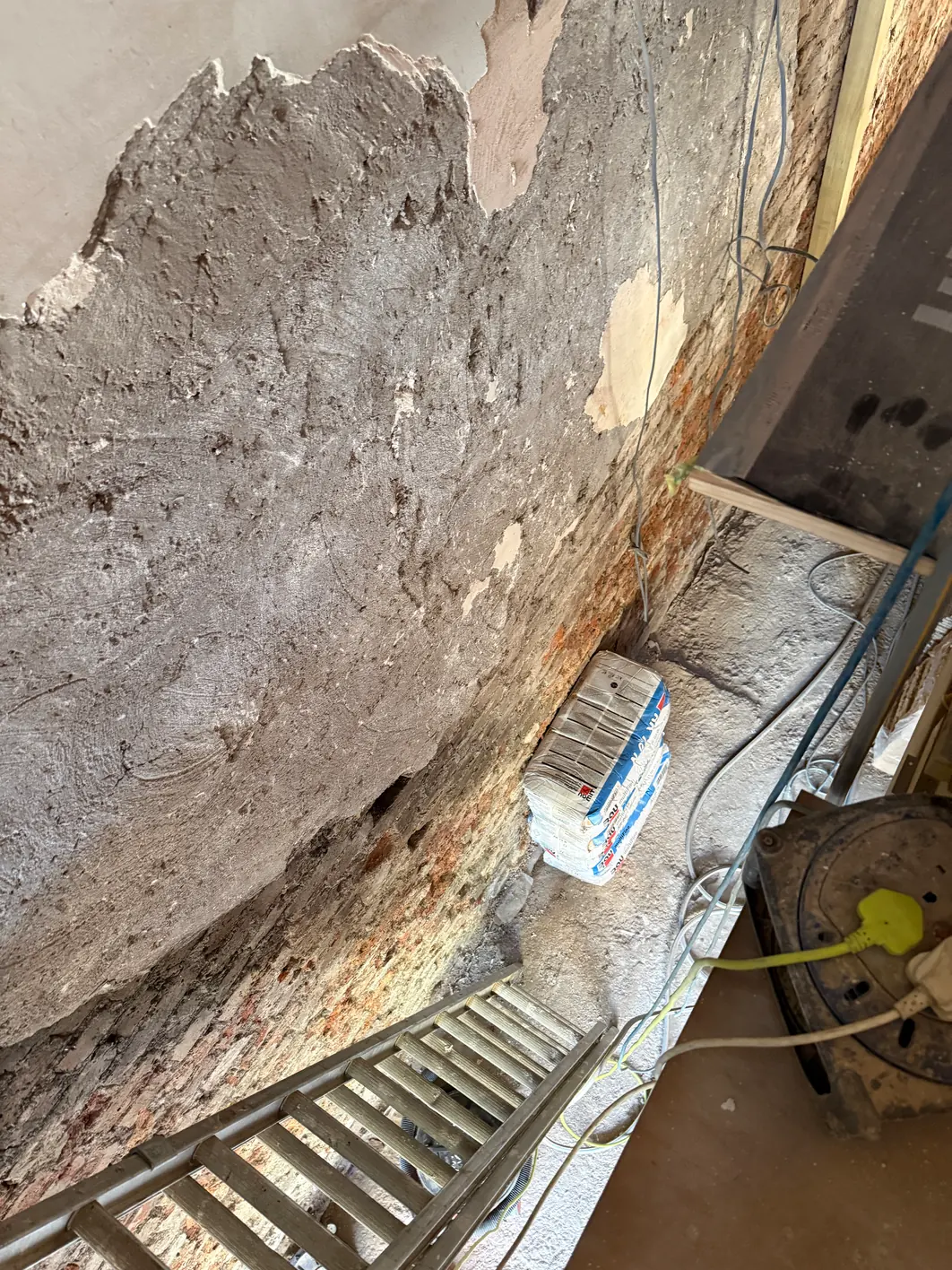 Wall stripped back to show a thick grey ash lime mortar coat, brick and stone showing through where it has gone, the original pale lime top coat above and a small pink patch of gypsum