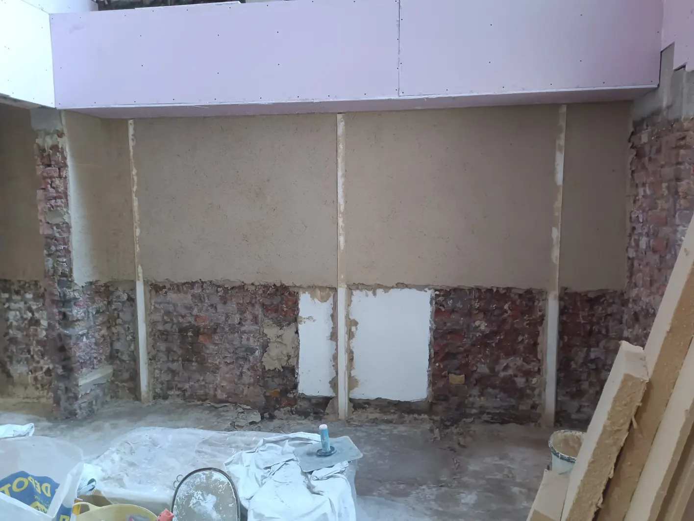 Wall taken back to bare brick with vertical plaster screeds set out along it and base coat filled in between them, brick still open along the lower half