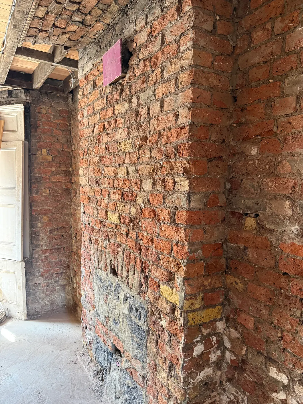 Lower ground floor room stripped to bare brick on every wall, mortar scars across the brick faces, a single small patch of pink plaster left high up
