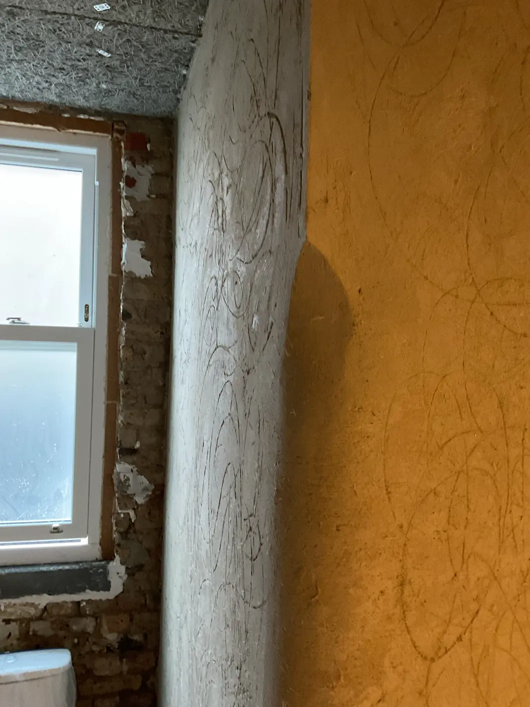 Window reveal and return in a pale float coat marked with looping devil-float scratches, beside an ochre wall marked the same way, with brick still exposed around the window