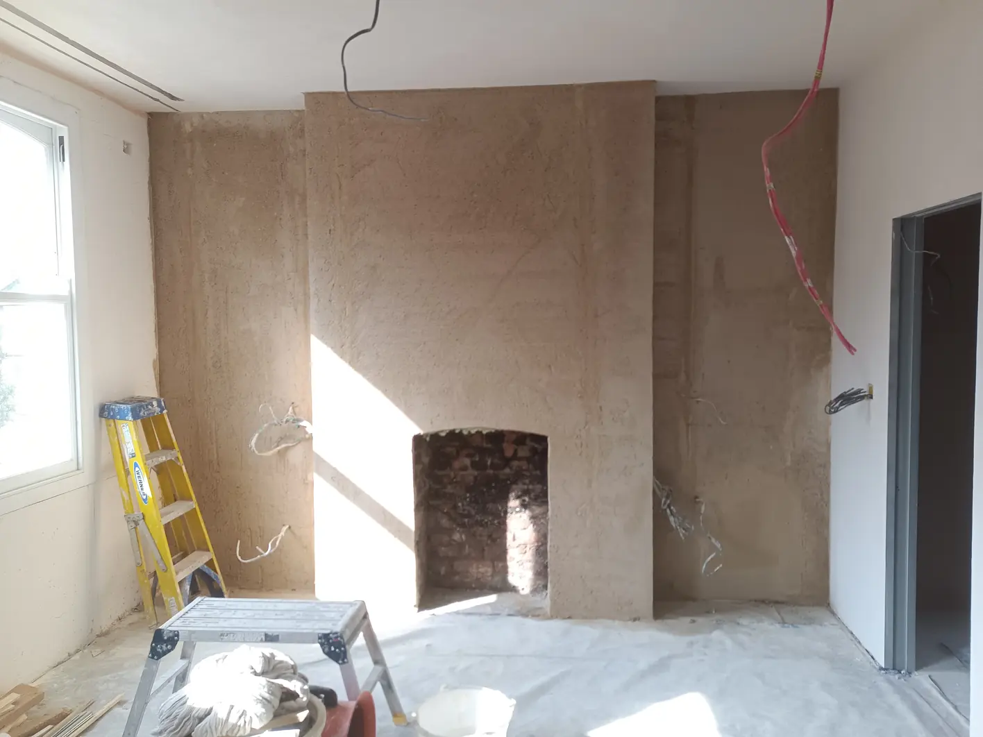 Chimney breast and both alcoves covered in an even base coat, the brick fireplace opening left exposed, window to the left and doorway to the right