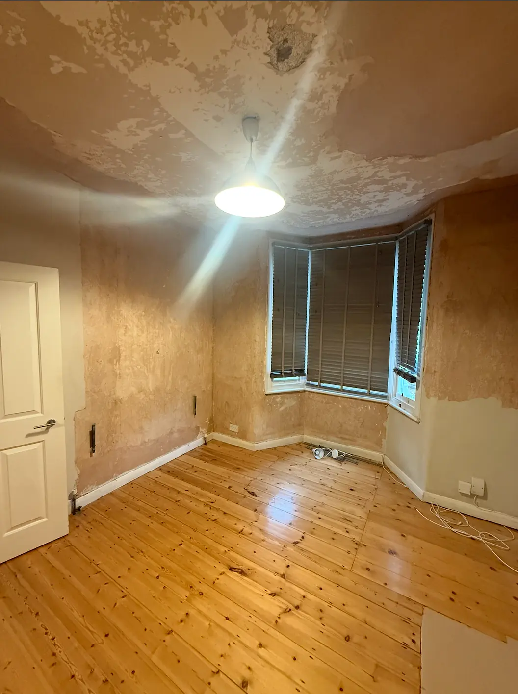 Empty room stripped ready for work, ceiling patchy where old finishes have come away, walls back to a bare undercoat, bare boards underfoot and a bay window with the blinds down