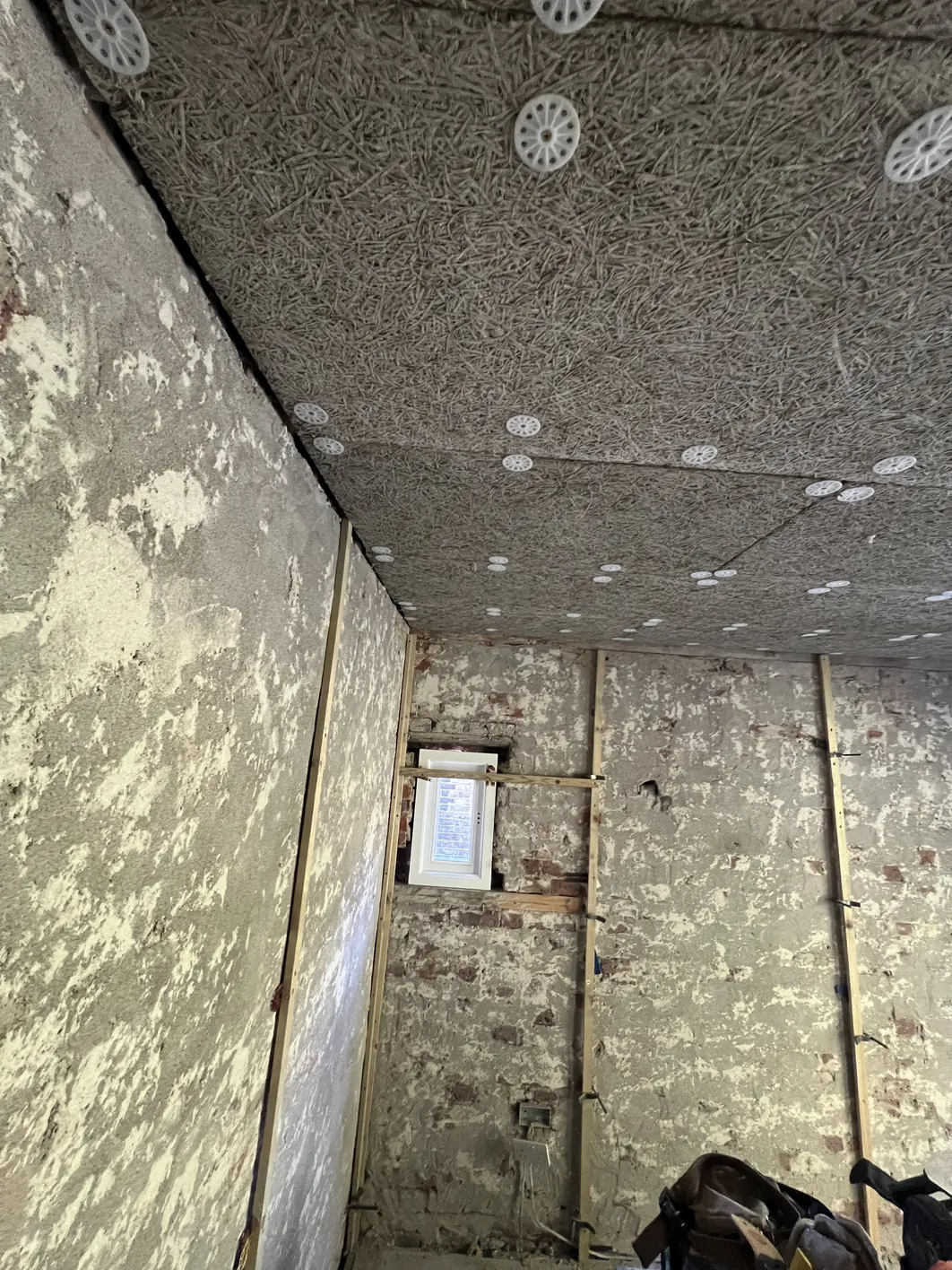 Overhead view of a ceiling showing grey wood wool board freshly installed with white ventilation sockets, mounted over deteriorated underlying plaster and brick walls.