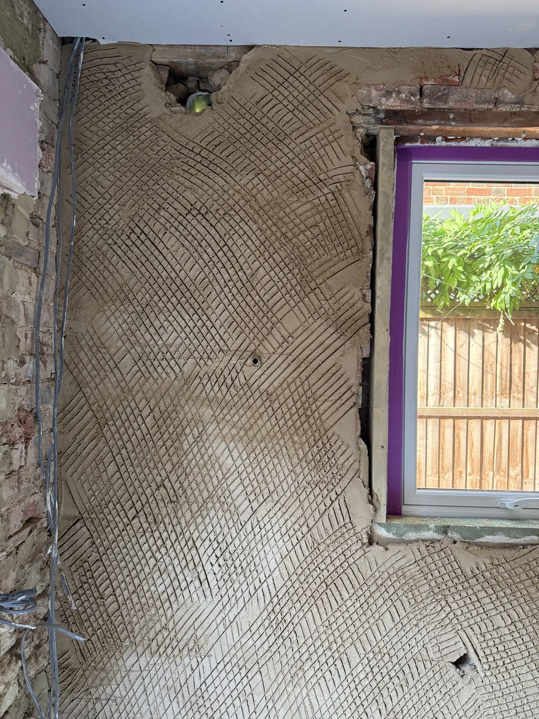 A mesh-reinforced plaster base coat with diagonal scoring pattern covers a solid brick wall beside a window with a purple frame.