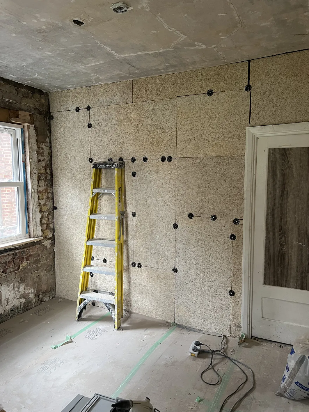 Interior room during construction showing tan-coloured insulation boards mechanically fixed to wall with black fixings, a yellow stepladder, plasterboard ceiling, window with exposed brick, and an open doorway.