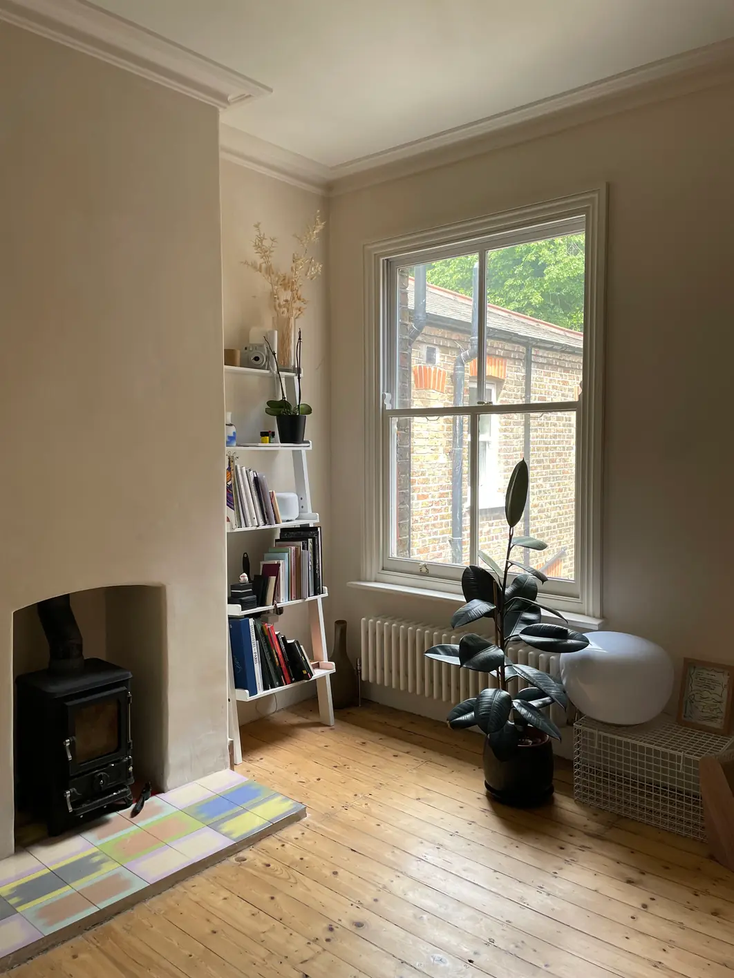 Living room with lime plaster walls meeting a retained cornice, a woodburning stove on a patterned tiled hearth, bookshelves and a large plant by the sash window