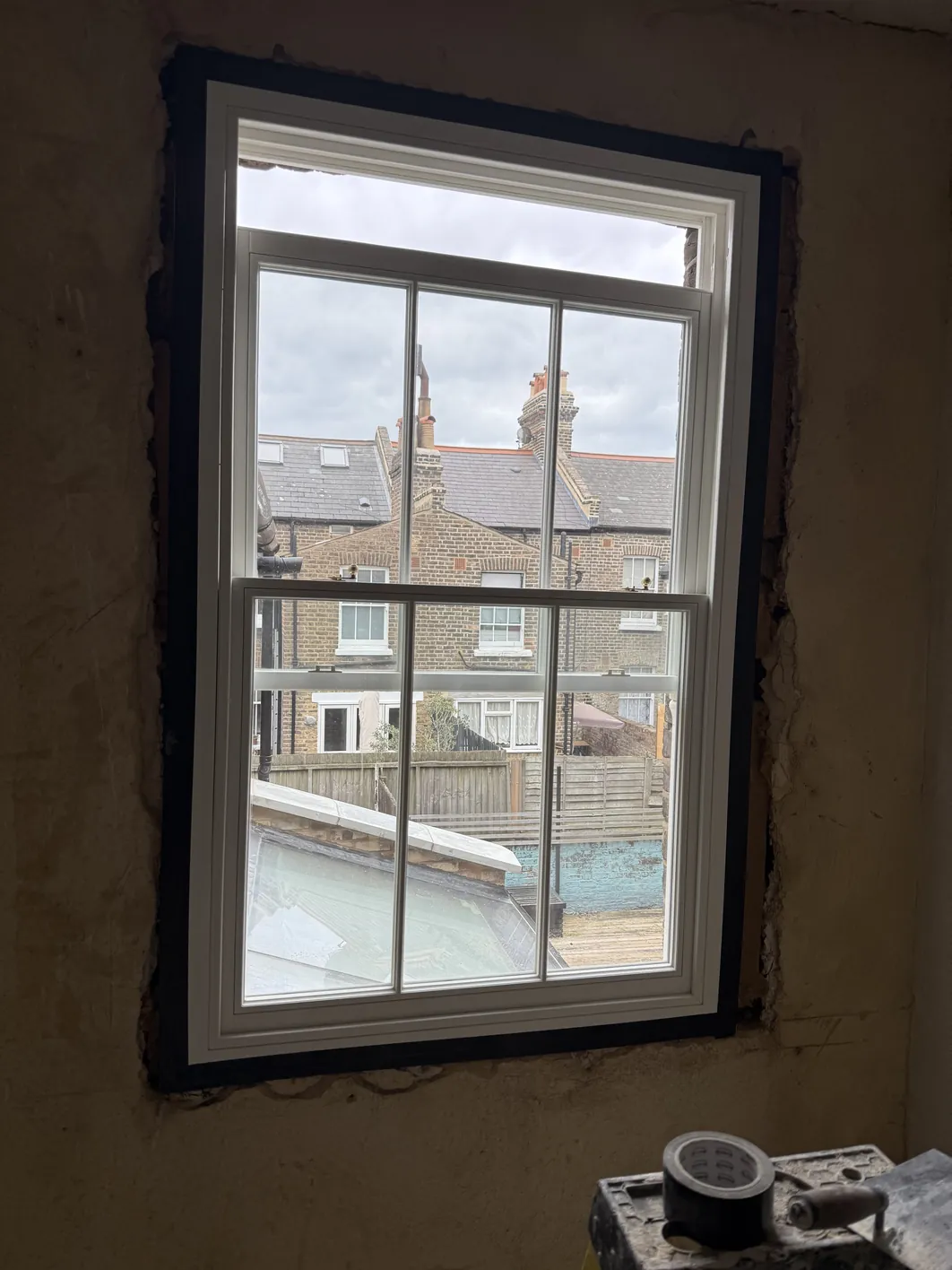 A new timber sash window with glazing bars and white frame installed in a wall cavity, viewed from inside during installation work.