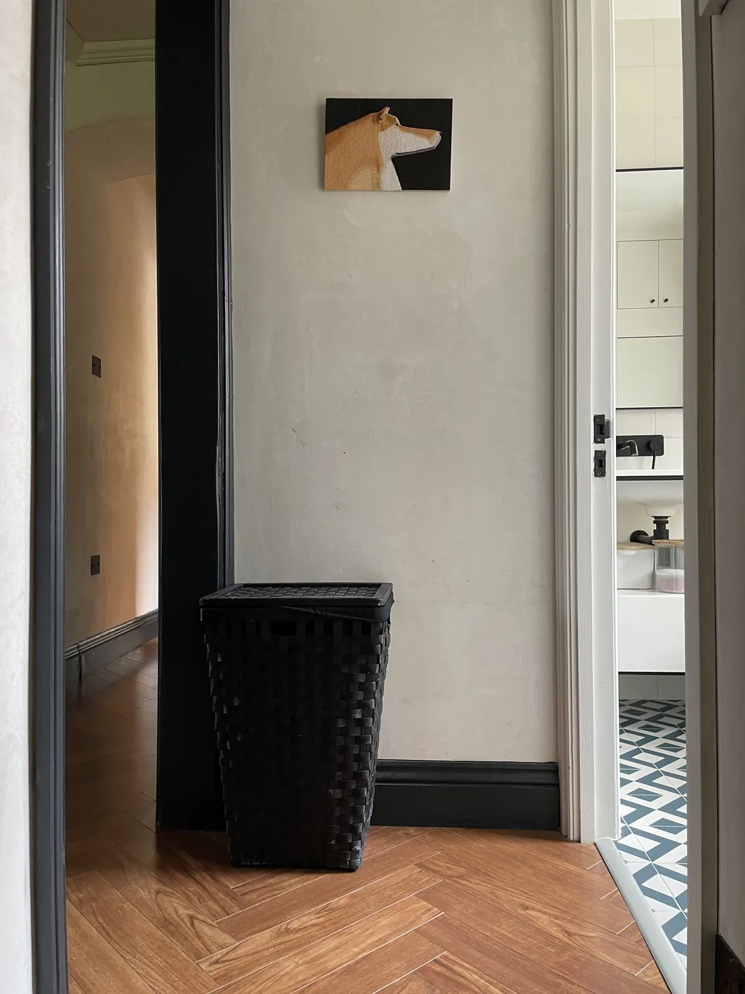 A light-filled hallway with plain cream-coloured plaster walls, a black wainscot baseboard, wooden herringbone floor, and open doorways on either side.