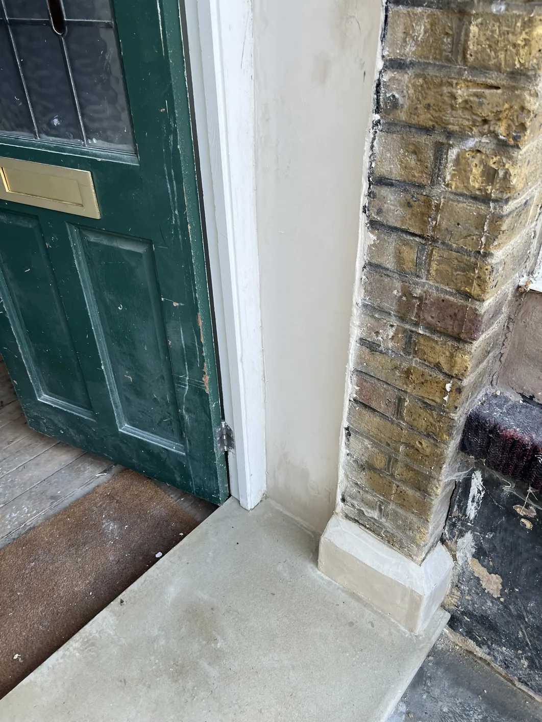 Interior doorway with green door on left, white-rendered wall and reveal in centre, and honey-coloured exposed brick wall on right with dark window sill below