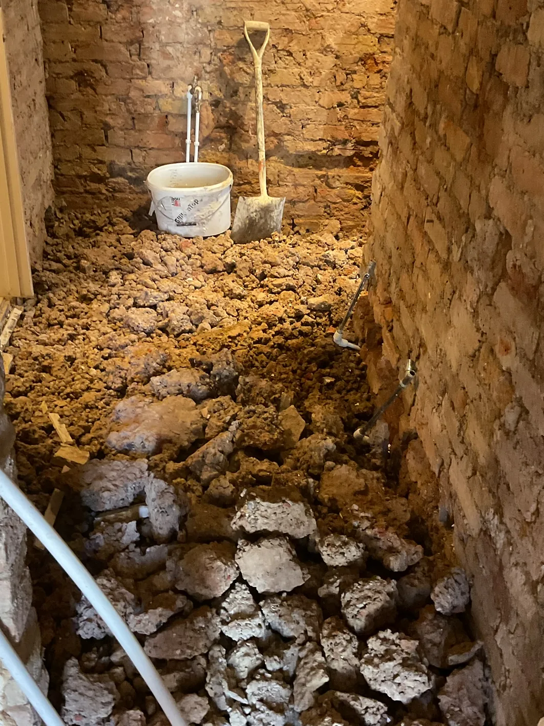 Excavated sub-floor void down to the brick footings, with a bucket and shovel left mid-dig, before any insulation goes in.