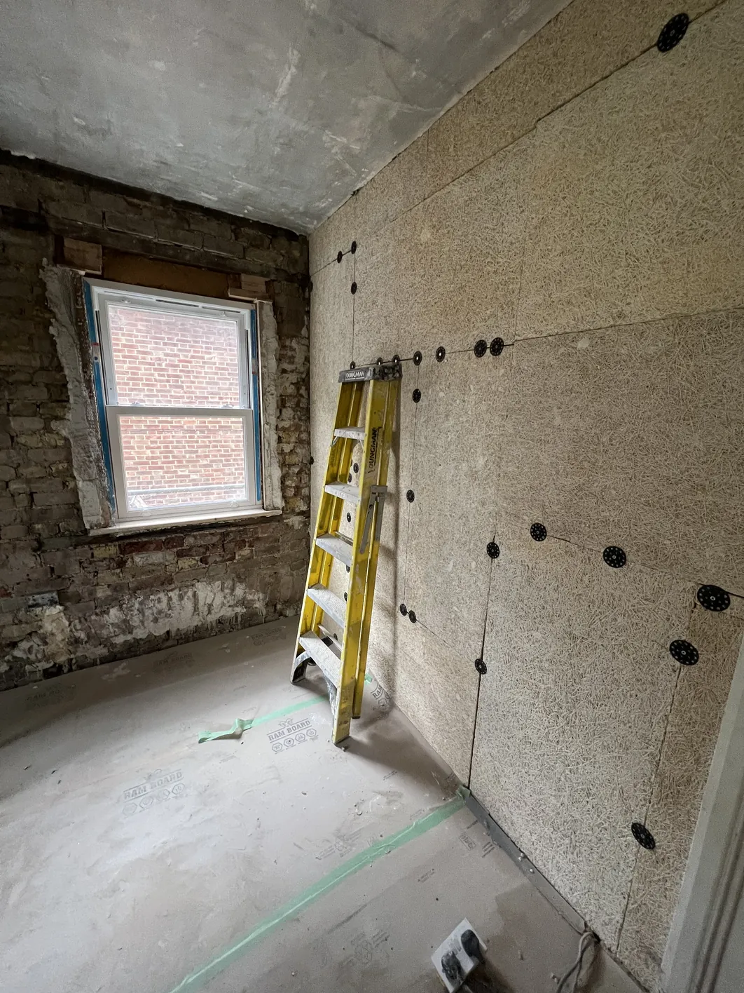 Interior room with exposed brick wall on left, wood fibre insulation board fixed to right wall with black fixings, window frame visible, step ladder in corner