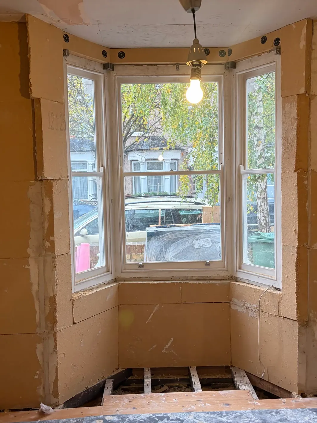 Bay window reveal lined on all three faces with wood fibre insulation board, fixing discs along the head, exposed floor joists below and a street view through the window