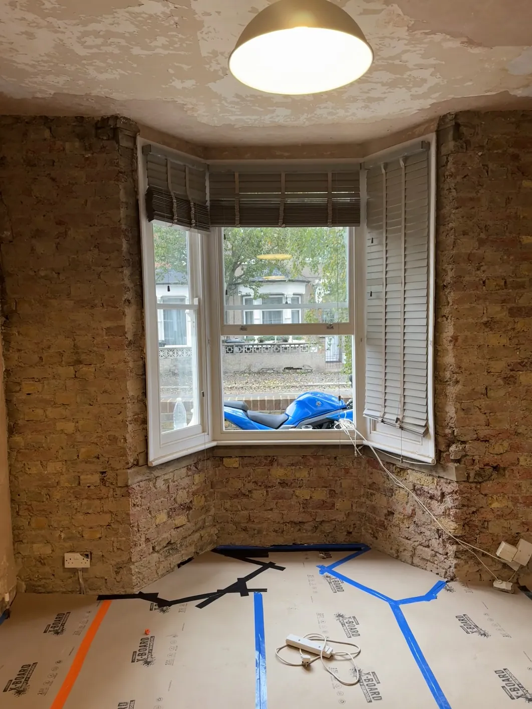 A bay window in a period property stripped to bare brick, with a new floor base layer and electrical conduits marked out in coloured tape