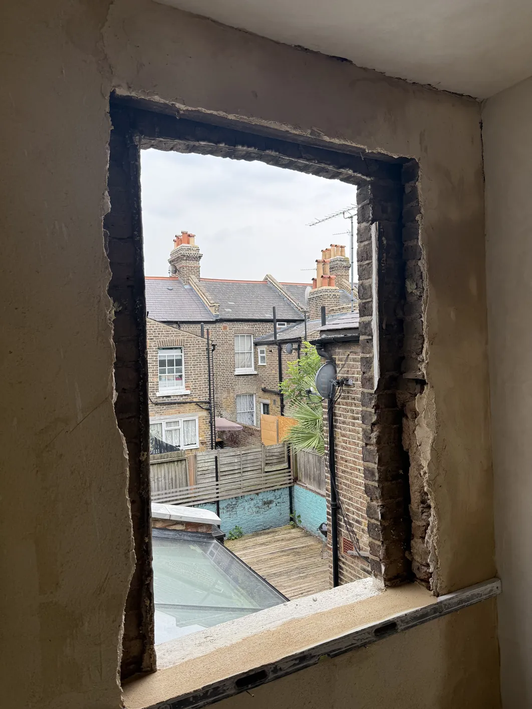 Interior view through a newly opened window reveal with dark rendered brick sides and a concrete lintel, opening onto a brick-paved courtyard with neighbouring period terraced houses.